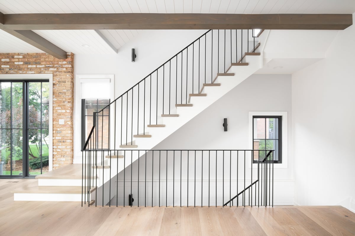 A beautiful staircase with wood flooring and stair treads, wrought iron railings, wood beams across the ceiling with shiplap, and a brick wall around a black framed window