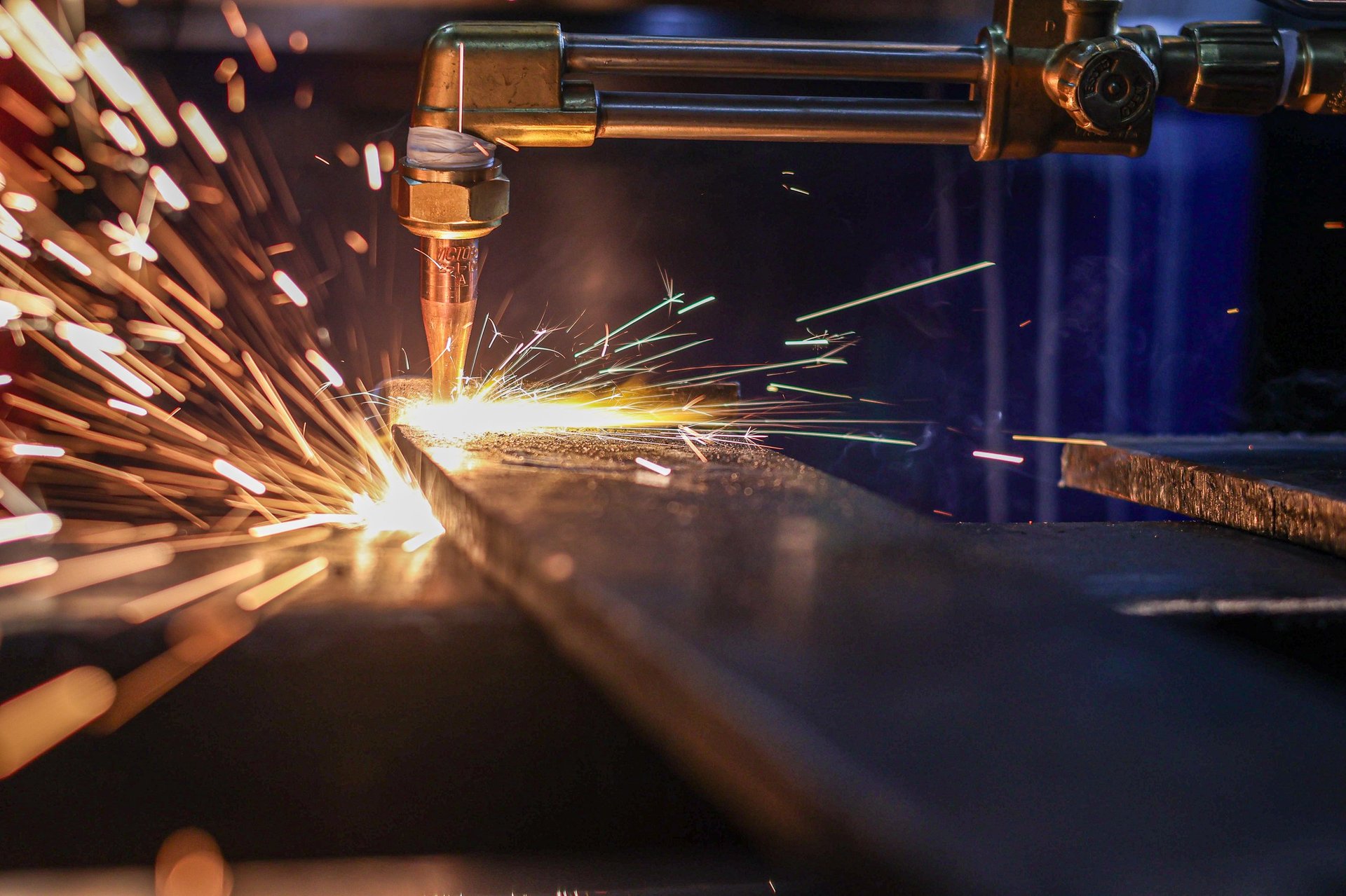 An industrial oxy-acetylene torch cuts through a thick steel plate, creating a dramatic spray of glowing sparks and molten metal in a fabrication workshop.