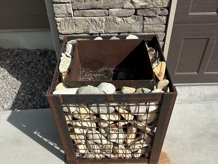 Rusty metal trash bin with metal mesh sides sitting on a concrete driveway next to a stone wall