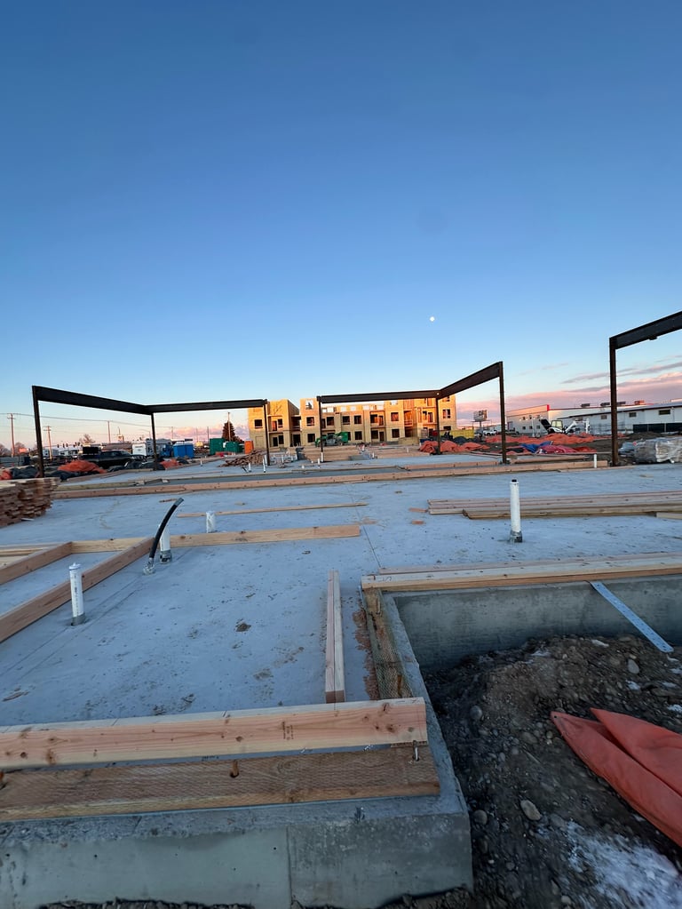 Construction site at dusk with concrete foundation, wooden framing, and a yellow building under construction in the background