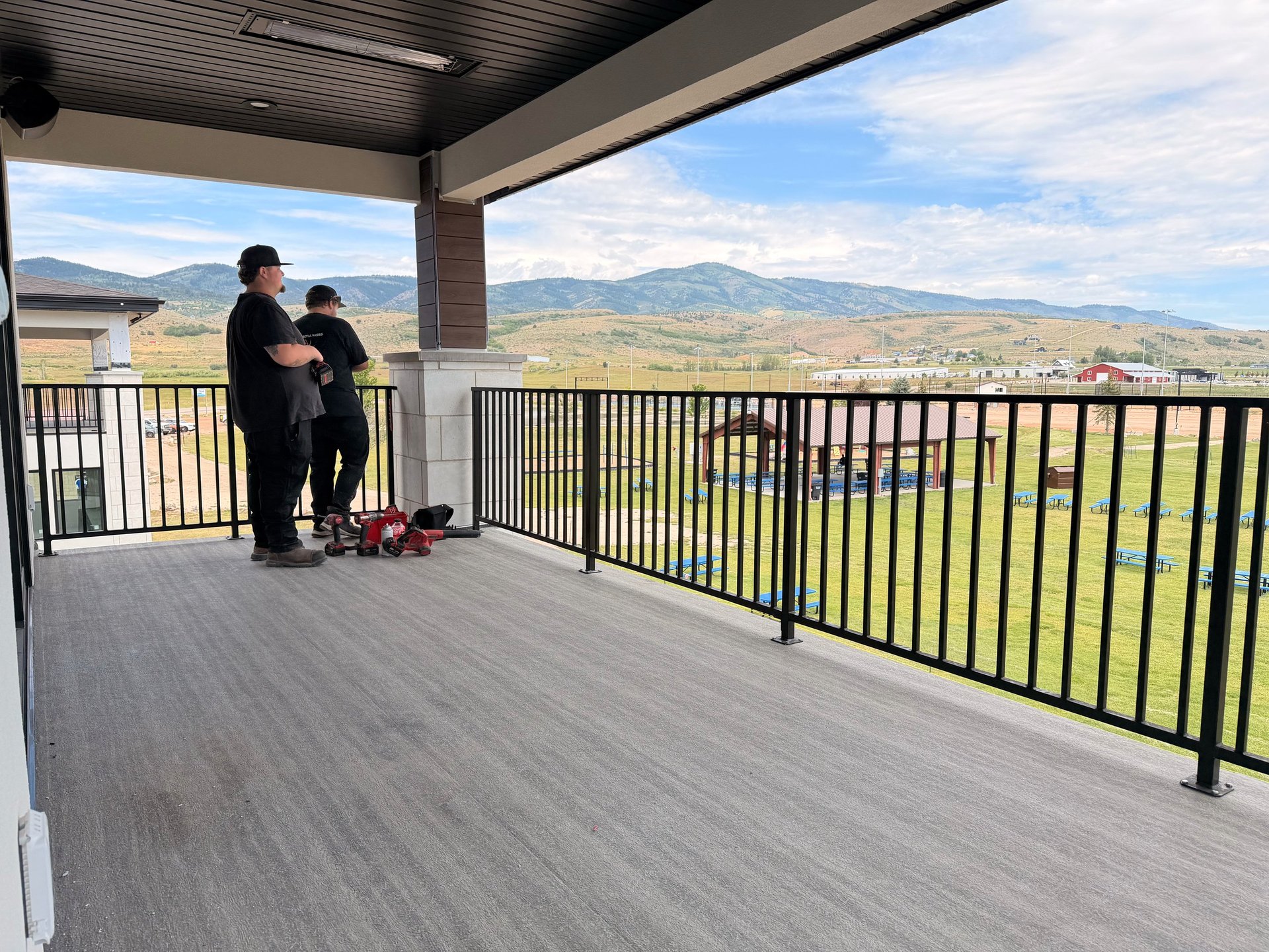 Two people standing on a spacious covered deck with black railing, overlooking a mountainous valley landscape