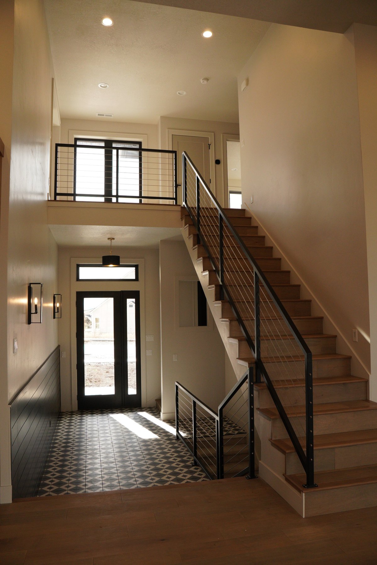 Modern foyer with wooden staircase, black metal railing, upper loft with glass door, and geometric tile floor entry