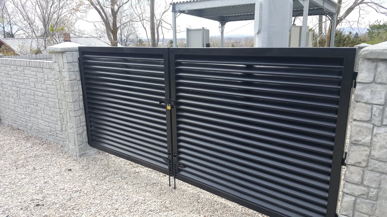 Black horizontal slatted metal driveway gate installed between white stone pillars on gravel surface