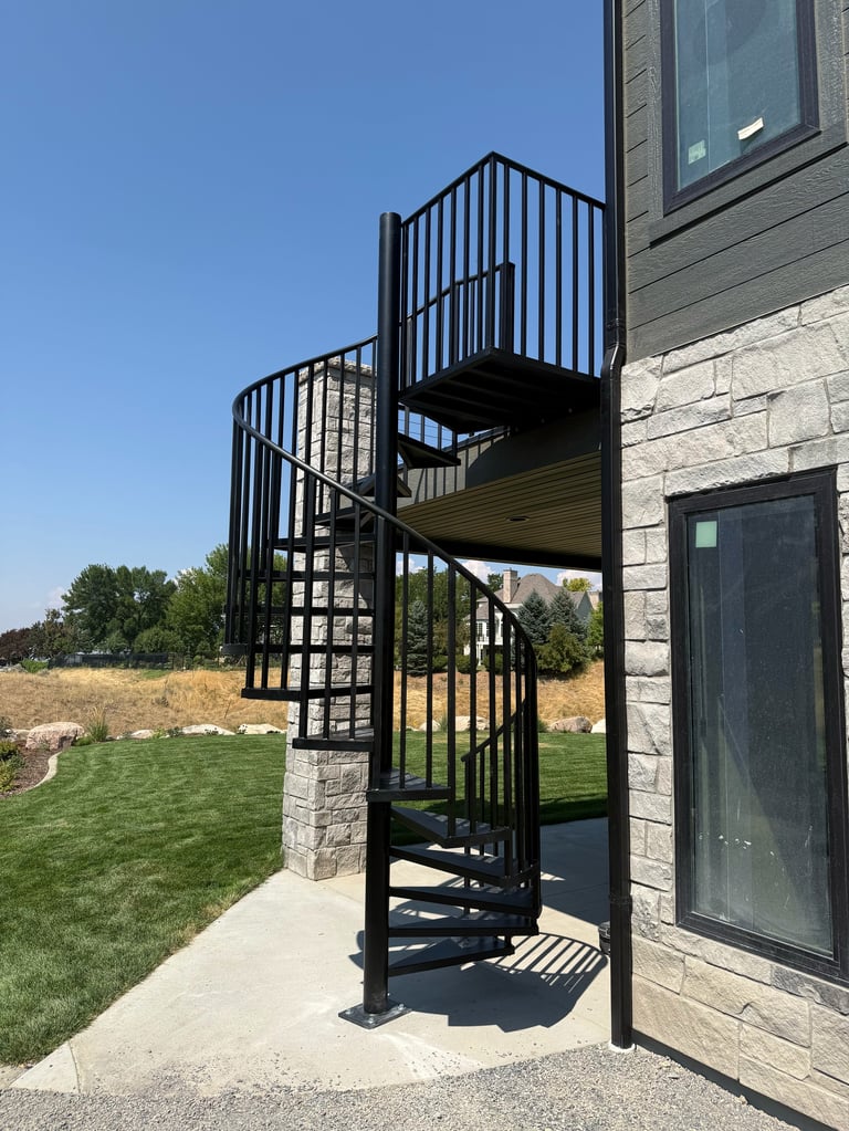 Black spiral staircase attached to stone house exterior with backyard and clear sky visible in background