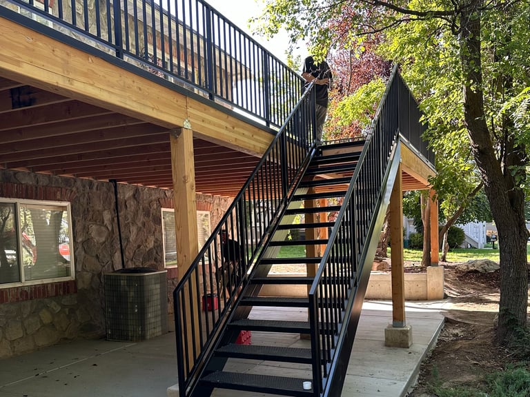 Modern wooden deck with metal railings and outdoor staircase leading to upper level, surrounded by trees