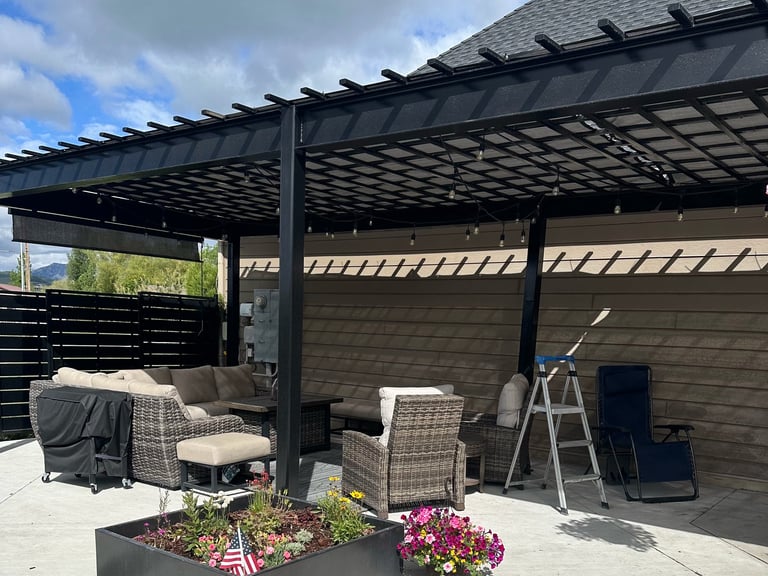 Modern black pergola with adjustable louvers over a patio seating area with wicker chairs, flowers, and outdoor furnishings