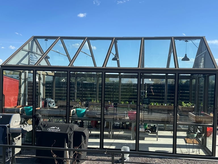 Modern black glass and metal greenhouse structure with geometric frame design against blue sky, containing garden plants and equipment inside