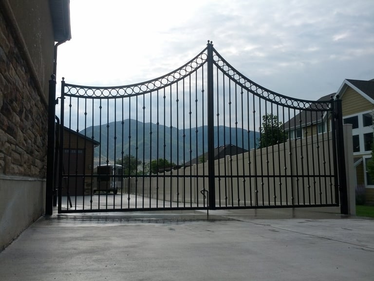 Ornate black iron driveway gate with decorative curved top, open on a paved entry with mountains visible beyond