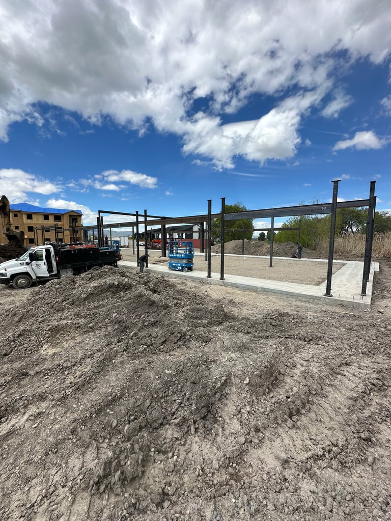 Construction site with metal frame structure, white pickup truck, and dirt ground under blue cloudy sky